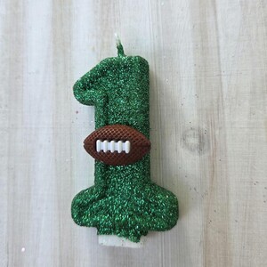 May include: A number one shaped candle covered in green glitter. A small brown football with white laces is attached to the center of the candle. The candle features a single white wick at the top and a flat white base at the bottom for stability.