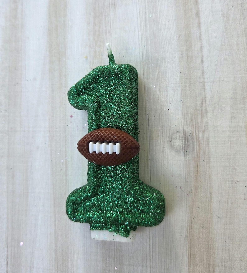 May include: A number one shaped candle covered in green glitter. A small brown football with white laces is attached to the center of the candle. The candle features a single white wick at the top and a flat white base at the bottom for stability.