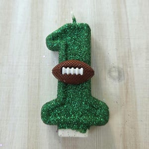 May include: A number one shaped birthday candle covered in green glitter. A small brown football with white laces is attached to the center of the candle. The candle has a white wick at the top and a white base at the bottom for stability.