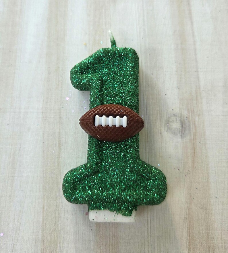May include: A number one shaped birthday candle covered in green glitter. A small brown football with white laces is attached to the center of the candle. The candle has a white wick at the top and a white base at the bottom for stability.
