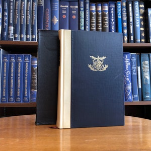 Può includere: Tre libri rilegati in tela con copertine blu scuro e dettagli dorati. I libri sono su un tavolo di legno. Il libro al centro ha uno stemma dorato sulla copertina.