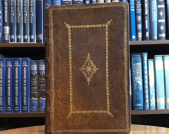 May include: A brown leather-bound book with gold accents and a decorative design on the front cover. The book is sitting on a wooden table.
