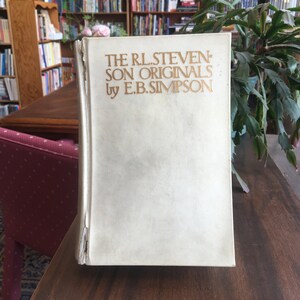 May include: A white book with gold lettering on the cover. The title is "The R.L. Stevenson Son Originals: by E.B. Simpson".