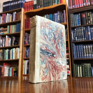 May include: A vintage book with a marbled cover in shades of blue, red, and white. The book's title is "TRACTS &c. BY THE LATE DR. DARBY". The book is sitting on a wooden surface in front of a bookshelf filled with books.