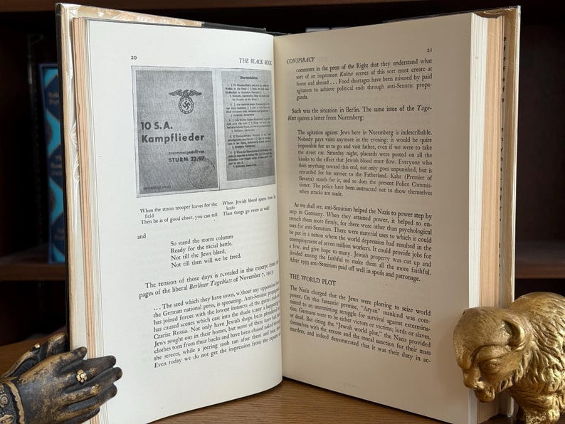 K&ouml;nnte beinhalten: Ein offenes Buch mit gealterten, vergilbten Seiten. Die linke Seite zeigt ein gedrucktes Bild mit deutschem Text. Die rechte Seite enth&auml;lt gedruckten Text. Ein dekorativer goldfarbener Bison-Buchst&uuml;tze ist sichtbar.