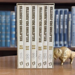 May include: A collection of six vintage books titled "RELATIONS DES JESUITES" in a white box, with dates from 1611 to 1672. A gold-coloured bison figurine is to the right of the books. The background shows a bookshelf with blue books.