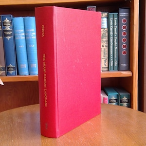 Op de afbeelding: Een rood hardback boek getiteld "The Miami-Illinois Language" door Costa, dat op een houten tafel ligt.