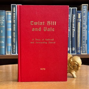 Könnte beinhalten: Ein rotes Buch mit dem Titel "Cwixt Hill and Vale" mit goldener Schrift, veröffentlicht im Jahr 1970. Das Buch steht aufrecht vor einem Regal mit anderen Büchern. Eine kleine goldene Büffelstatue steht auf dem Tisch.
