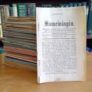 May include: A stack of old, bound books with a single book on top titled "Sameiningin" in black lettering. The book is open to a page with text in a foreign language. The book is on a wooden table in front of a bookshelf.