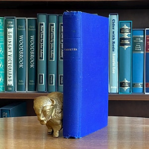 Może przedstawiać: Niebieska książka w twardej oprawie zatytułowana "Vanity Fair" z imieniem autora nadrukowanym złotymi literami. Książka znajduje się na drewnianej półce z innymi książkami.