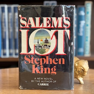 Könnte beinhalten: Ein gebundenes Exemplar von Stephen Kings Roman "Salem's Lot" mit schwarzem Einband und roter und weißer Schrift. Das Cover zeigt eine Illustration einer Stadt. Das Buch steht auf einer Holzoberfläche.