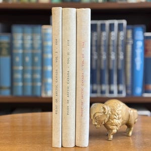May include: Three vintage books titled "Pilot of Arctic Canada" with the years 1959 and 1961. A gold-colored bison figurine sits to the right of the books. The books are on a wooden surface with a bookshelf in the background.