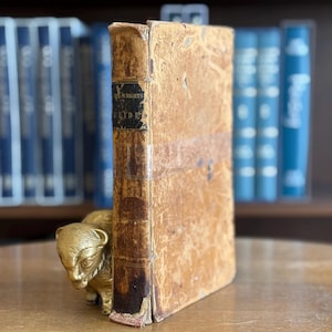 Puede incluir: Un libro antiguo con una cubierta de cuero curtido desgastado se encuentra en posición vertical, junto a una figura de bisonte dorada. El lomo del libro tiene una etiqueta negra con texto descolorido. Libros azules son visibles en el fondo, sugiriendo una biblioteca o estudio.