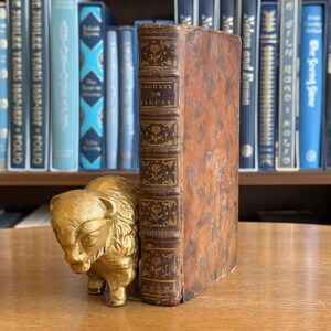 May include: A vintage book with a worn, brown leather cover and gold embossed spine, titled "RECUEIL DE PIECES." A gold-colored bison figurine stands beside the book on a wooden surface, with a backdrop of antique books on a shelf.