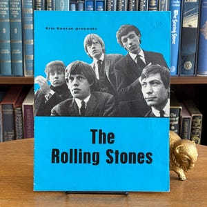 Könnte beinhalten: Ein Vintage-Albumcover für The Rolling Stones mit einem Schwarz-Weiß-Foto der Bandmitglieder vor einem hellblauen Hintergrund. Der Bandname steht in weißen, fetten Buchstaben. Präsentiert von Eric Easton.