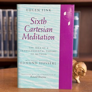 Puede incluir: Un libro titulado "Sixth Cartesian Meditation" de Eugen Fink. La portada es azul claro con texto morado y un lomo morado. El libro está sobre una superficie de madera. El texto del libro incluye el nombre de Edmund Husserl.