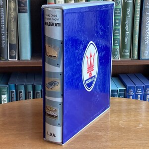Puede incluir: Un libro de tapa dura azul con el título "MASERATI" en letras blancas. El libro tiene un borde blanco y un logotipo de tridente plateado con la palabra "MASERATI" debajo. El libro está sobre una mesa de madera frente a una estantería.