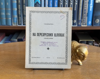 Puede incluir: Un libro antiguo titulado "HA ПЕРЕХРЕСНИХ ШЛЯХАХ" con texto en ucraniano, publicado en Canadá en 1954. El libro tiene una cubierta de color claro con bordes decorativos y está apoyado en un soporte. A la derecha hay una pequeña figura de bisonte dorada.