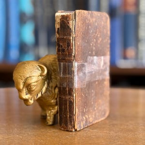 May include: An antique book with a distressed, brown leather cover is supported by a gold-coloured bison figurine. The book's spine is taped with clear tape. The background is blurred, showing a bookshelf with blue books.