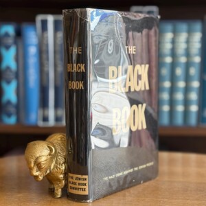 K&ouml;nnte beinhalten: Ein gebundenes Buch mit dem Titel "The Black Book" mit goldfarbenen Buchstaben. Der Einband des Buches hat einen schwarzen Hintergrund mit einem schwarz-wei&szlig;en Bild. Vor dem Buch steht eine kleine goldene B&uuml;ffelfigur.