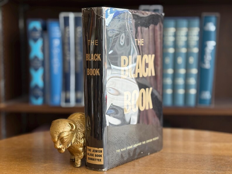K&ouml;nnte beinhalten: Ein gebundenes Buch mit dem Titel "The Black Book" mit goldfarbenen Buchstaben. Der Einband des Buches hat einen schwarzen Hintergrund mit einem schwarz-wei&szlig;en Bild. Vor dem Buch steht eine kleine goldene B&uuml;ffelfigur.