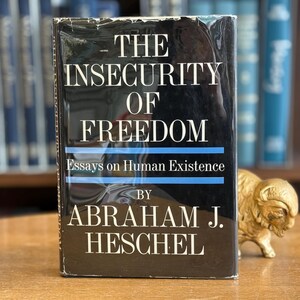 Könnte beinhalten: Ein Buch mit dem Titel "The Insecurity of Freedom" von Abraham J. Heschel. Das Buch hat einen schwarzen Einband mit weißem Text und blauen Linien. Neben dem Buch steht eine kleine goldfarbene Bisonfigur auf einer Holzoberfläche. Essays über die menschliche Existenz.