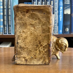 May include: A vintage bookend with a weathered, tan leather cover. The bookend is supported by a gold-colored bison sculpture. The background features a bookshelf with various antique books.