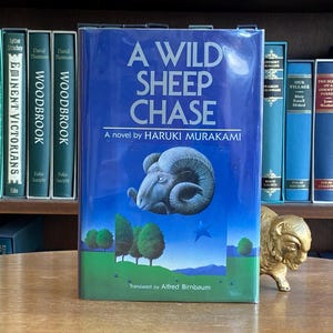 Może przedstawiać: Niebieska książka w twardej oprawie zatytułowana "A Wild Sheep Chase" autorstwa Haruki Murakami. Okładka książki przedstawia kreskówkową owcę z dużymi rogami i gwiazdą.