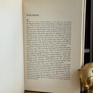 K&ouml;nnte beinhalten: Aufgeschlagenes Buch mit dem Titel "Indictment" auf der linken Seite. Der Text behandelt die Geschichte der Juden in Europa. Eine goldfarbene B&auml;renfigur liegt auf der rechten Seite. Das Buch befindet sich in einer Bibliothek.