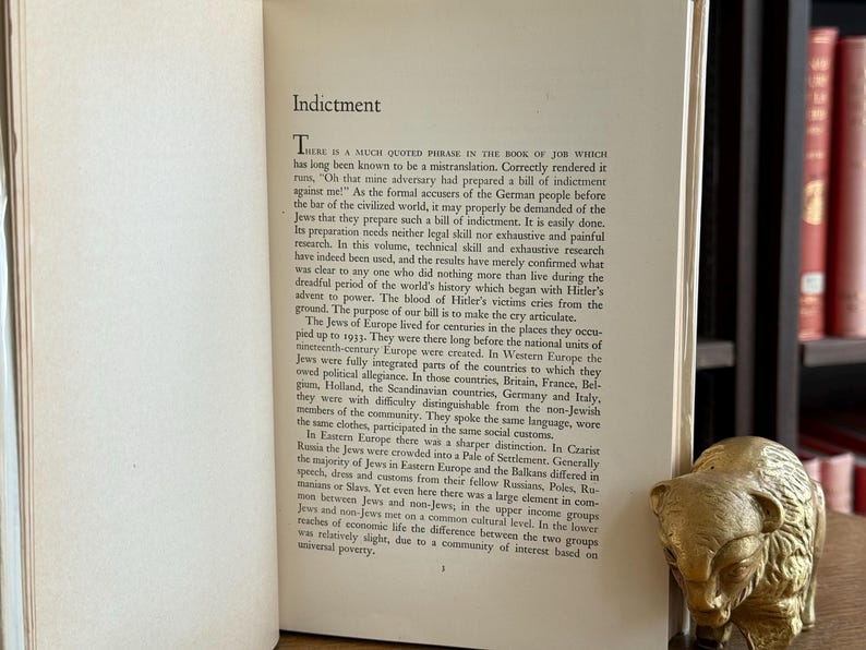 K&ouml;nnte beinhalten: Aufgeschlagenes Buch mit dem Titel "Indictment" auf der linken Seite. Der Text behandelt die Geschichte der Juden in Europa. Eine goldfarbene B&auml;renfigur liegt auf der rechten Seite. Das Buch befindet sich in einer Bibliothek.