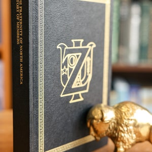 May include: A black leather-bound book with gold lettering and a gold embossed crest. The book is titled "Zeta Psi Fraternity of North America Directory of Members" and the year 1987 is printed on the bottom. The book is sitting on a wooden surface.