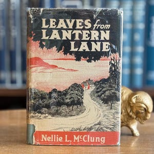 Puede incluir: Un libro antiguo titulado "Leaves from Lantern Lane" con una ilustración de portada de un camino que conduce a una casa. El libro tiene una cubierta negra con detalles en rojo y blanco. El nombre del autor, Nellie L. McClung, está impreso en la parte inferior.