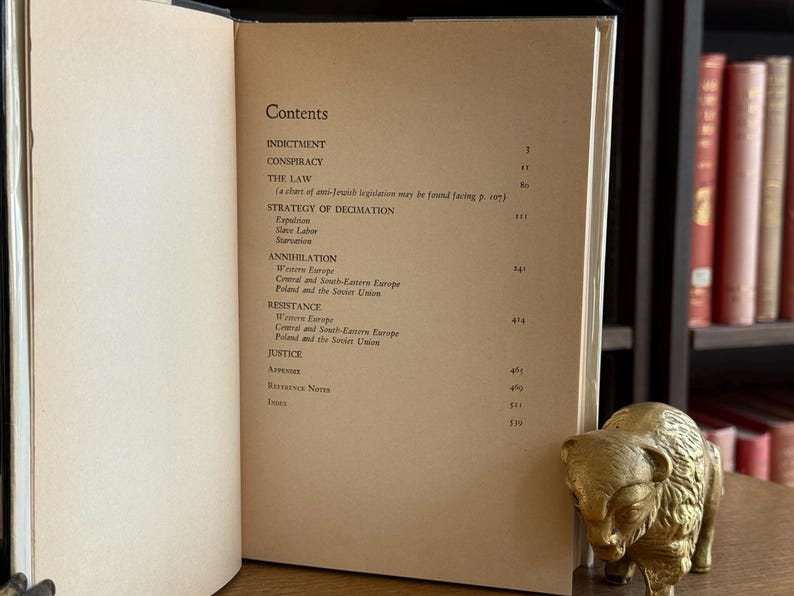 K&ouml;nnte beinhalten: Aufgeschlagenes Buch mit Inhaltsverzeichnis, das Titel wie &bdquo;Indictment&ldquo; und &bdquo;Conspiracy&ldquo; enth&auml;lt. Eine kleine, goldfarbene Bisonfigur liegt auf dem Tisch. B&uuml;cherregale sind im Hintergrund zu sehen.
