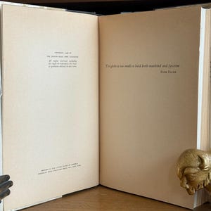 K&ouml;nnte beinhalten: Ein aufgeschlagenes Buch mit beigefarbenen Seiten, auf denen Text gedruckt ist. Auf der rechten Seite befindet sich eine goldfarbene Bison-Statue. Der Buchr&uuml;cken ist sichtbar, und die Seiten wirken leicht gealtert.
