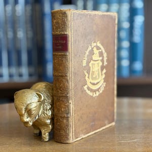 Puede incluir: Un libro antiguo con una cubierta de cuero marrón y letras doradas, sujeto por una figura de búfalo dorada. El lomo del libro tiene una etiqueta roja con texto. El fondo muestra una estantería borrosa.