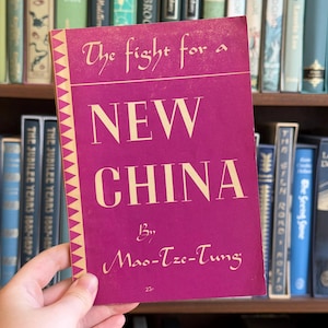 Könnte beinhalten: Ein Vintage-Buch mit dem Titel "The Fight for a New China" von Mao-Tze-Tung. Das Buch hat einen magentafarbenen Einband mit weißem Text und einem dekorativen gelb-weißen Rand. Das Buch wird vor einem Bücherregal mit anderen Büchern gehalten.