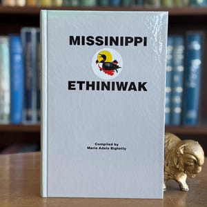 May include: A hardcover book titled "MISSINIPPI ETHINIWAK" with a circular emblem featuring a bird. The book is light gray with black text and is compiled by Marie Adele Bighetty. A small gold buffalo statue is on the right.