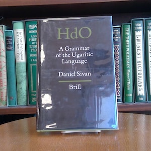 May include: A dark gray hardcover book titled "HdO: A Grammar of the Ugaritic Language" by Daniel Sivan. The book is published by Brill and is sitting on a wooden table in front of a bookshelf.