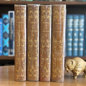 Puede incluir: Cuatro libros antiguos con cubiertas de cuero marrón y patrones decorativos dorados. Las lomos dicen "LEM.DESACY SAINTE BIBLE" con los números de volumen 1-4. Una figura de bisonte dorada está a la derecha. Otros libros azules están en el fondo.