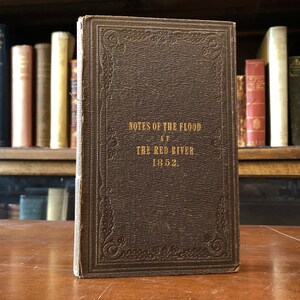 May include: A brown leather-bound book with gold lettering on the cover. The title is "Notes of the Flood at the Red River 1852."