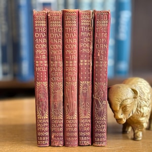 Puede incluir: Una colección de libros antiguos con cubiertas rojas y letras doradas, incluyendo títulos como "Dante" y "La Divina Comedia". Una figura dorada de búfalo está cerca. Los libros están apilados verticalmente.
