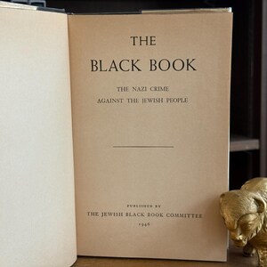 K&ouml;nnte beinhalten: Ein offenes Buch mit dem Titel "The Black Book" mit Text &uuml;ber die Verbrechen der Nazis an den Juden, ver&ouml;ffentlicht 1946. Rechts vom Buch befindet sich eine goldfarbene Bisonfigur.