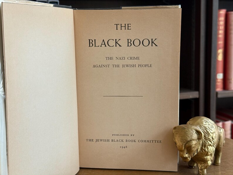K&ouml;nnte beinhalten: Ein offenes Buch mit dem Titel "The Black Book" mit Text &uuml;ber die Verbrechen der Nazis an den Juden, ver&ouml;ffentlicht 1946. Rechts vom Buch befindet sich eine goldfarbene Bisonfigur.