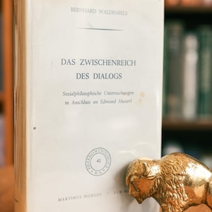 Pode incluir: Um livro branco com o título "Das Zwischenreich des Dialogs" de Bernhard Waldenfels. O livro faz parte de uma série chamada "Phaenomenologica" e é o número 41 da série. O livro está sobre uma mesa de madeira com uma figura de búfalo de metal dourado ao lado.