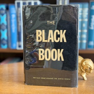 K&ouml;nnte beinhalten: Ein schwarzes Hardcover-Buch mit dem Titel "THE BLACK BOOK" in goldener Schrift. Der Untertitel lautet "THE NAZI CRIME AGAINST THE JEWISH PEOPLE". Das Buch liegt auf einem Holztisch, im Hintergrund sind weitere B&uuml;cher zu sehen.