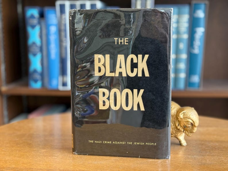 K&ouml;nnte beinhalten: Ein schwarzes Hardcover-Buch mit dem Titel "THE BLACK BOOK" in goldener Schrift. Der Untertitel lautet "THE NAZI CRIME AGAINST THE JEWISH PEOPLE". Das Buch liegt auf einem Holztisch, im Hintergrund sind weitere B&uuml;cher zu sehen.