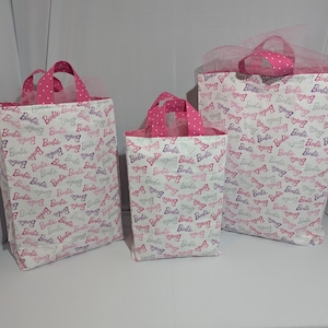 Peut inclure: Trois sacs cadeaux en tissu de différentes tailles. Les sacs sont blancs avec un motif répété du mot "Barbie" en rose et violet. Les poignées et la doublure intérieure sont roses avec des pois blancs. Le plus grand sac a une garniture en tulle rose.