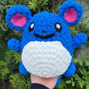 May include: A blue and white crocheted plush toy resembling a Pokemon character. The toy has large ears with pink insides and a smiling face.