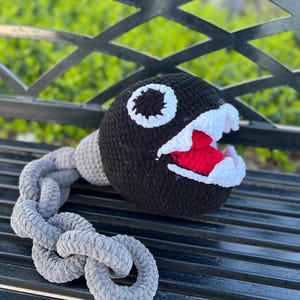 Może przedstawiać: Pluszowa zabawka w kształcie postaci z gry Super Mario Bros., wykonana na szydełku w kolorze czarnym i białym. Zabawka ma szeroko otwarte usta z czerwonymi zębami i długi, szary ogon wykonany na szydełku.