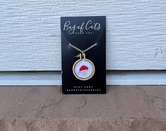 May include: A silver necklace with a small, embroidered mushroom pendant. The pendant is set in a miniature embroidery hoop. The necklace is displayed on a black card with the text "Bag of Cats" and "Thank You".
